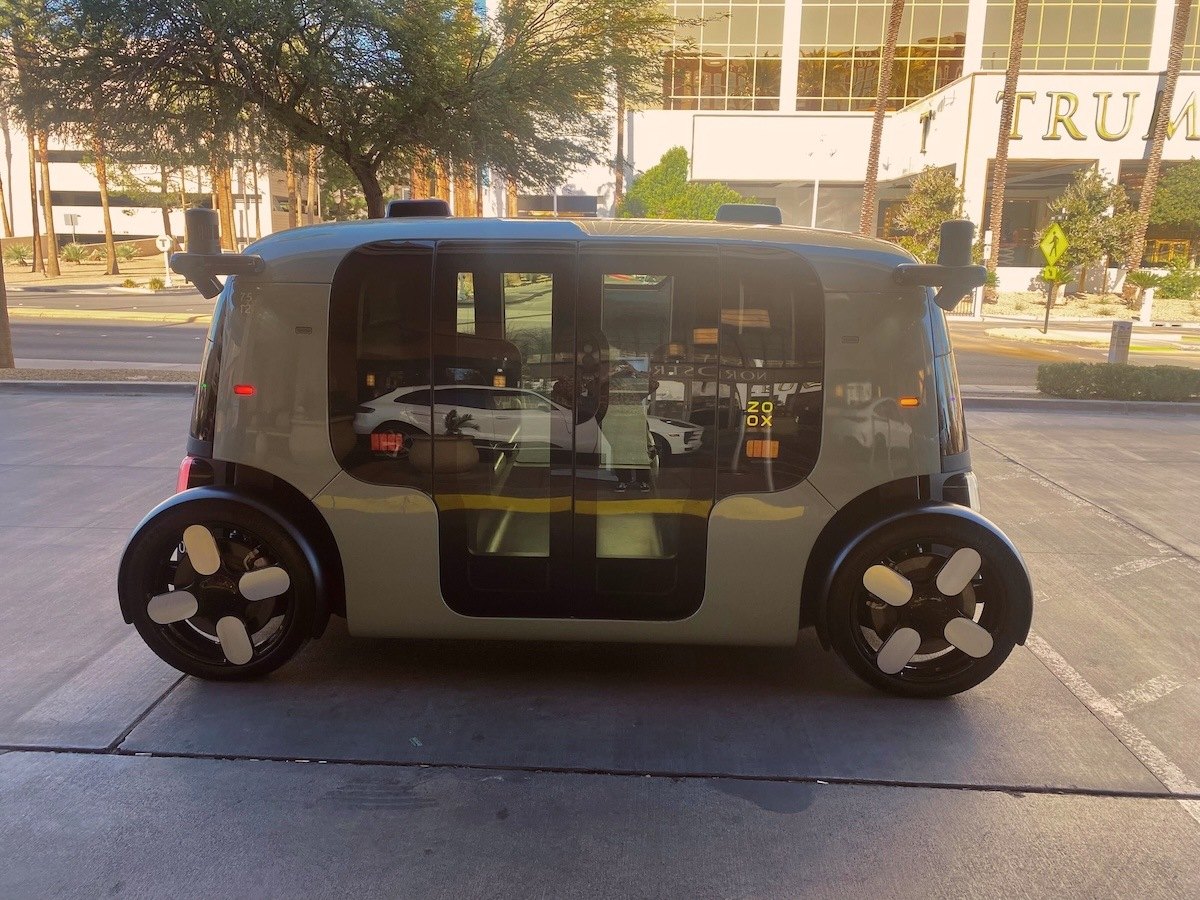 A Zoox autonomous ride-share vehicle parked at the rideshare pickup area outside Fashion Show Mall. The box-shaped car has large windows, sliding doors, and four wheels, with Trump International Hotel and trees visible in the background.